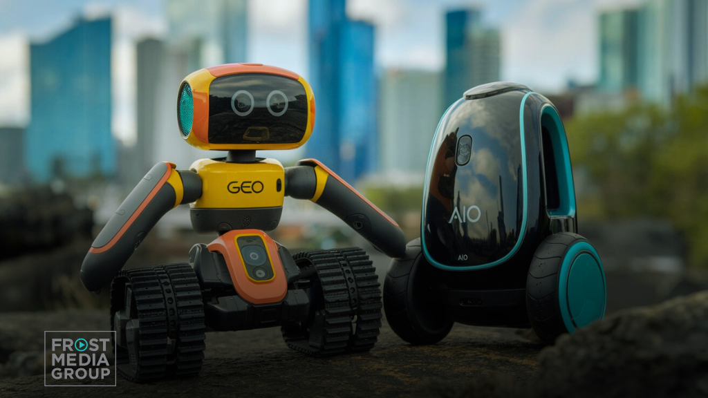 This image shows two small robots set against a blurred cityscape background. On the left, a yellow and orange robot with tank-like treads and "GEO" labeled on its chest has a humanoid design with arms and a digital face. To the right is a sleek, black-and-teal robot labeled "AIO," featuring a more rounded design on wheels, giving it a futuristic look. The Frost Media Group logo appears in the lower left corner. The scene portrays an innovative, technology-driven environment with the robots representing advancements in AI and robotics. GEO AIO