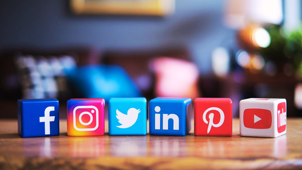 A photo of cute, colorful cubes with the logos of various social media platforms. There are cubes with the Facebook "f", Instagram "camera", Twitter "bird", LinkedIn "connect", Pinterest "pin", and YouTube "play button". The cubes are arranged in a row on a wooden surface. The background is blurred and consists of a living room with a couch, a lamp, and a plant.