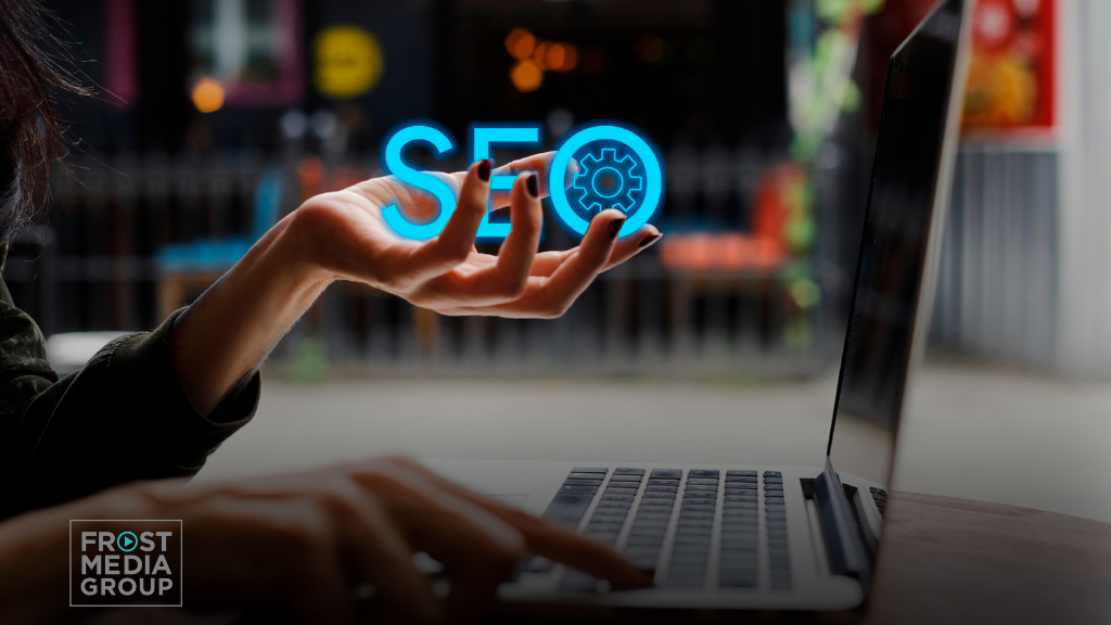 A close-up of a person using a laptop, with one hand hovering above the keyboard. The hand holds a glowing, blue 3D holographic icon of the letters 'SEO' alongside a gear symbol, symbolizing search engine optimization. The background is blurred, focusing attention on the hand and the holographic SEO graphic, creating a modern and technological feel. The image conveys the concept of digital marketing and optimization through advanced tools and techniques. Semantic SEO